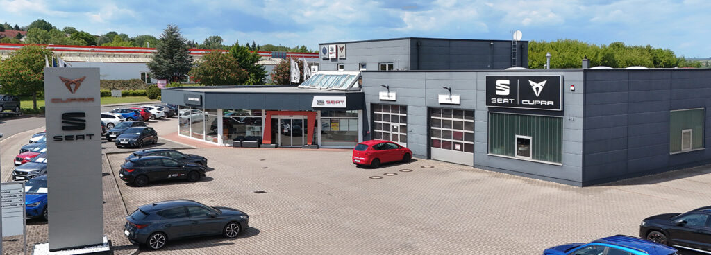 Nach der Überschrift ist eine Aussenaufnahne vom Autohaus Eichner in Eisleben zu sehen Im Vordergrund rechts an der Autohaus Einfahrt ein Pylon, mit den Logos Cupra und Seat. Rechts in der Mitte ist der Eingangsbereich des Autohaus. Hier finden Sie ihre Ansprechpartner des Servicebereichs und unser Verkaufsteam. Links im Bild der Werkstattbereich des Autohau Eichner.